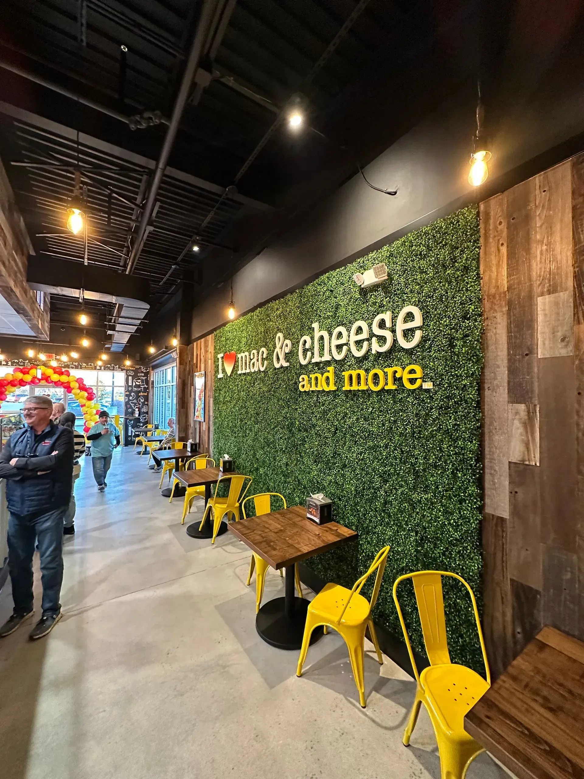 An indoor seating area with bright yellow metal chairs, wooden tables, and a green wall featuring the sign