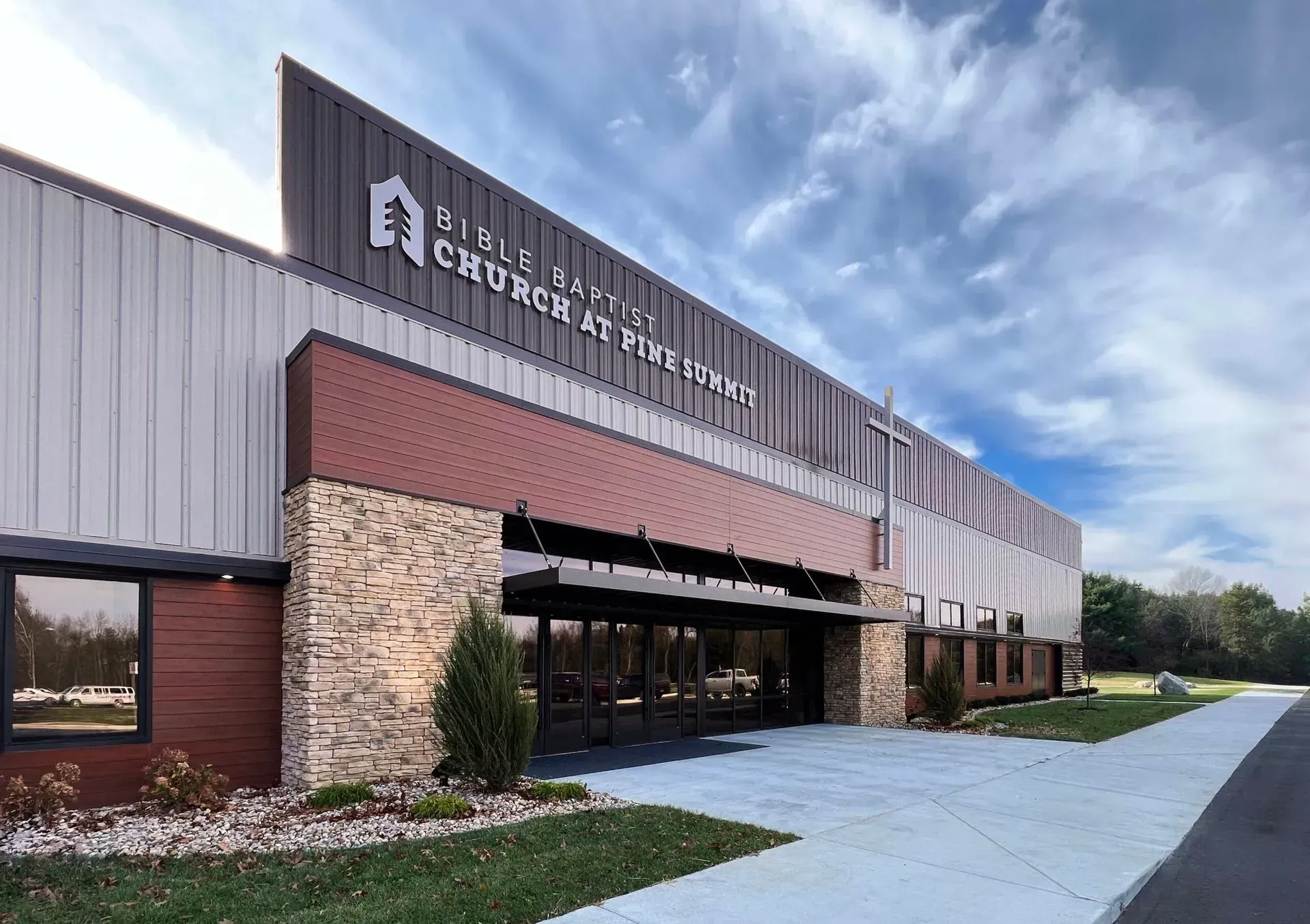 Modern church building with stone facade, glass entrance, metal siding, and a logo sign under a blue, partly cloudy sky.