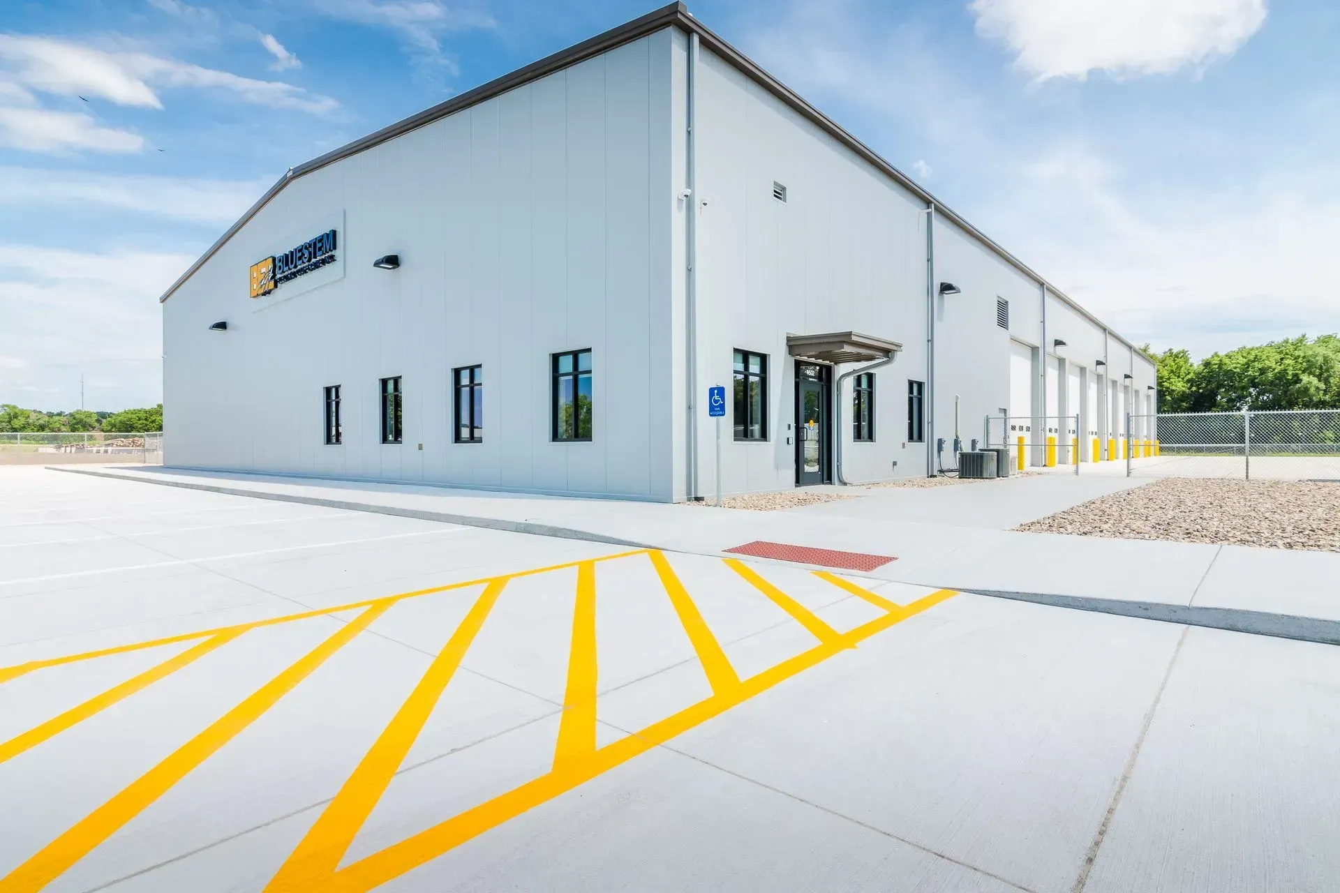A grey industrial warehouse building with a white exterior, multiple windows, and yellow painted markings on the asphalt.