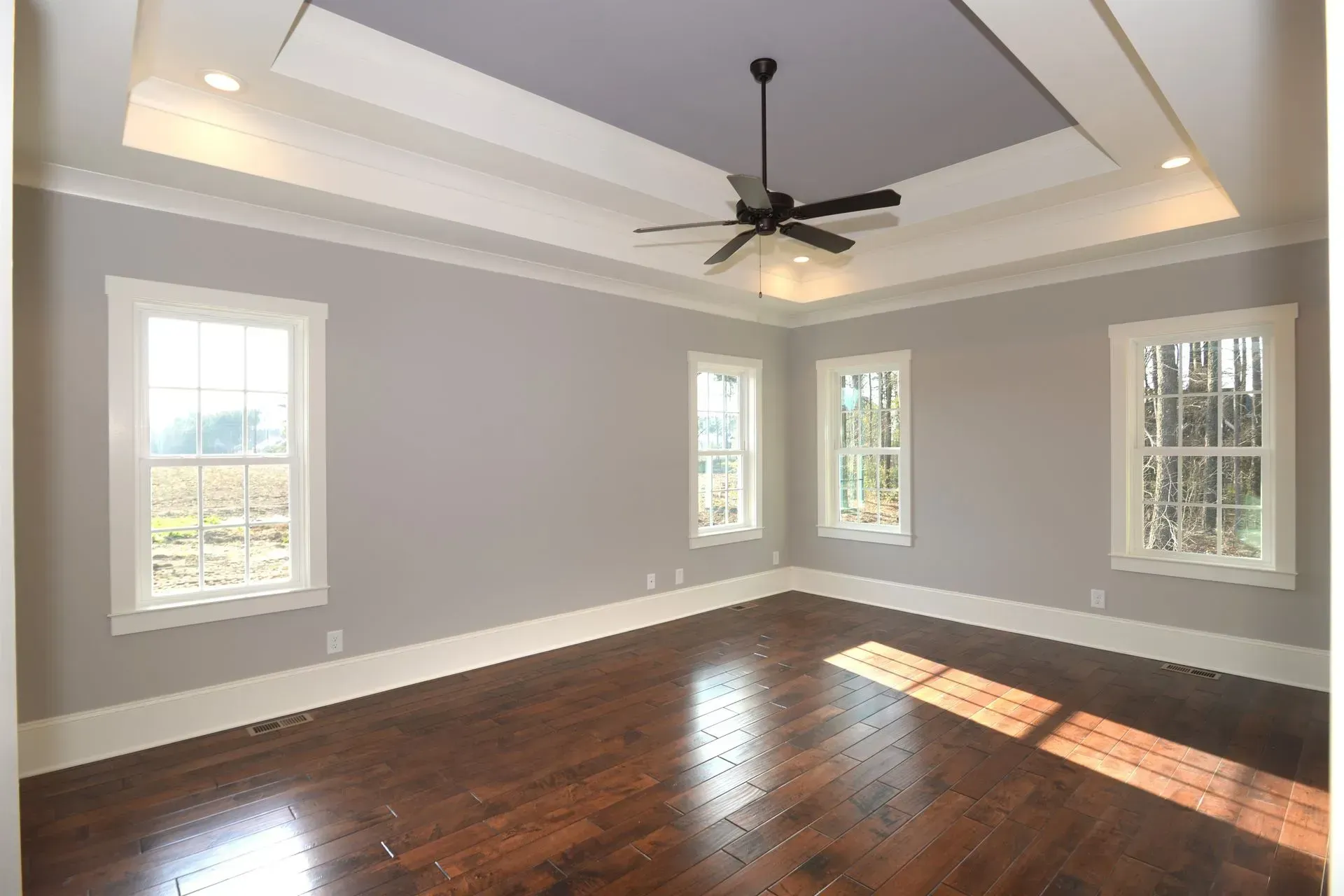 An empty bedroom with gray walls, white trim, a dark wood floor, and a tray ceiling with a fan and recessed lighting.