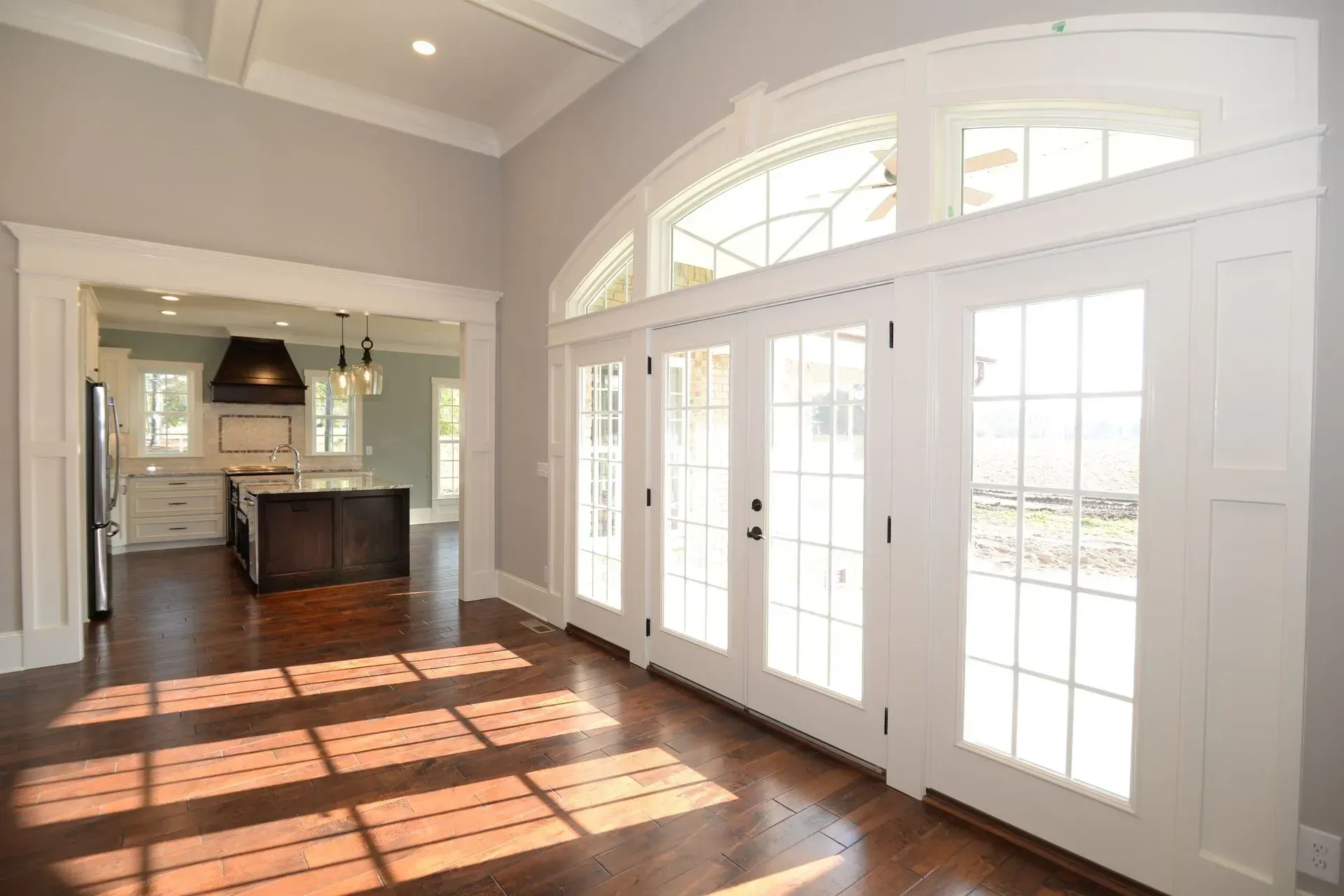 A wide, bright room with dark hardwood floors, a white arched glass door, and a view into a kitchen with an island.
