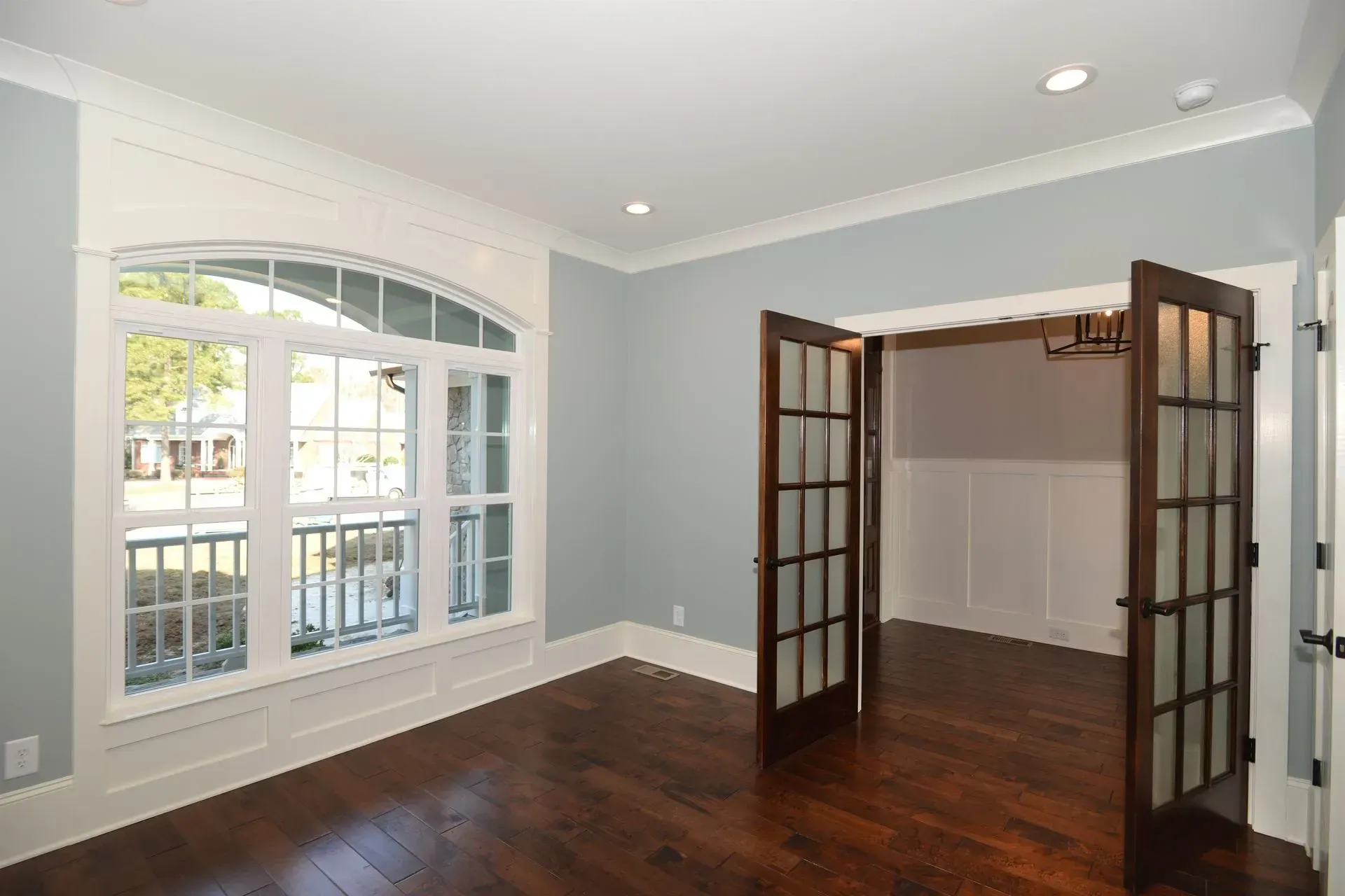 An empty room with light blue walls, dark hardwood floors, a large arched window, and open dark wood French doors.