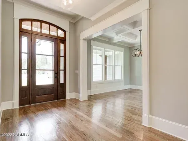 A light-filled foyer with hardwood floors, a large arched wooden front door, and a view into a bright room with tray ceilings.
