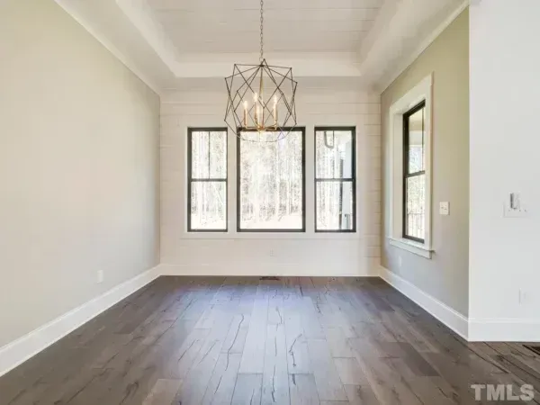A bright, empty dining room with gray wood floors, neutral walls, three large windows, and a modern geometric chandelier.