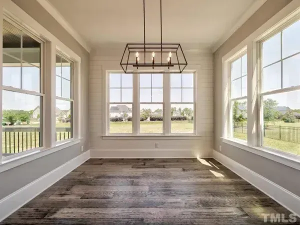 An empty sunroom with white shiplap walls, dark wood-look floors, large windows, and a rectangular black metal chandelier.