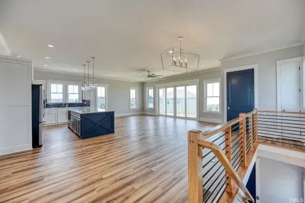 An open-concept living area with light wood floors, a dark blue kitchen island, and a wooden staircase in the foreground.