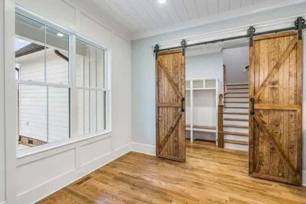 A room with hardwood floors, a large window, and double wooden barn doors opening to a hallway with stairs and cubbies.