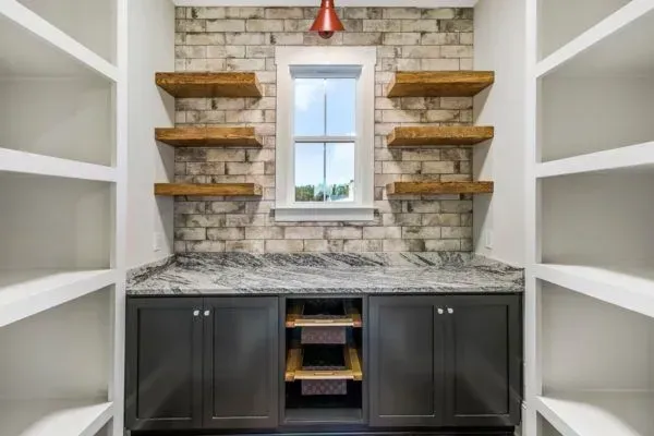 A pantry with dark cabinets, a stone countertop, open wooden shelves, and a window against a light brick wall.