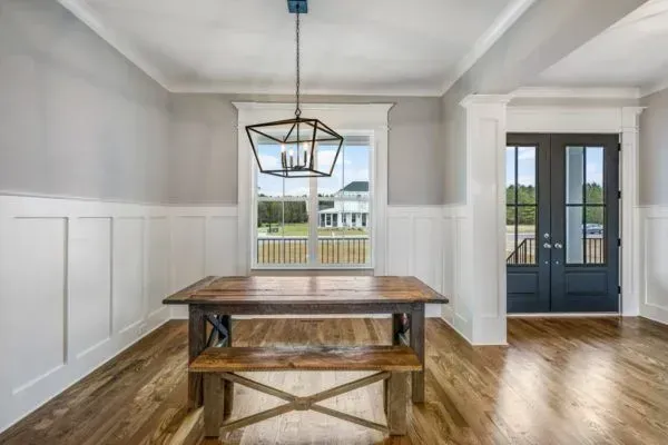 Dining room with a rustic wooden table, bench, geometric pendant light, white wainscoting, and dark double doors.