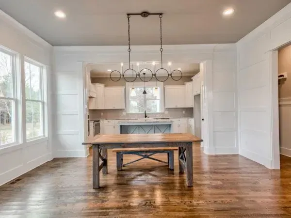 A rustic wooden dining table sits in a bright, modern dining room with hardwood floors and a white kitchen in the back.