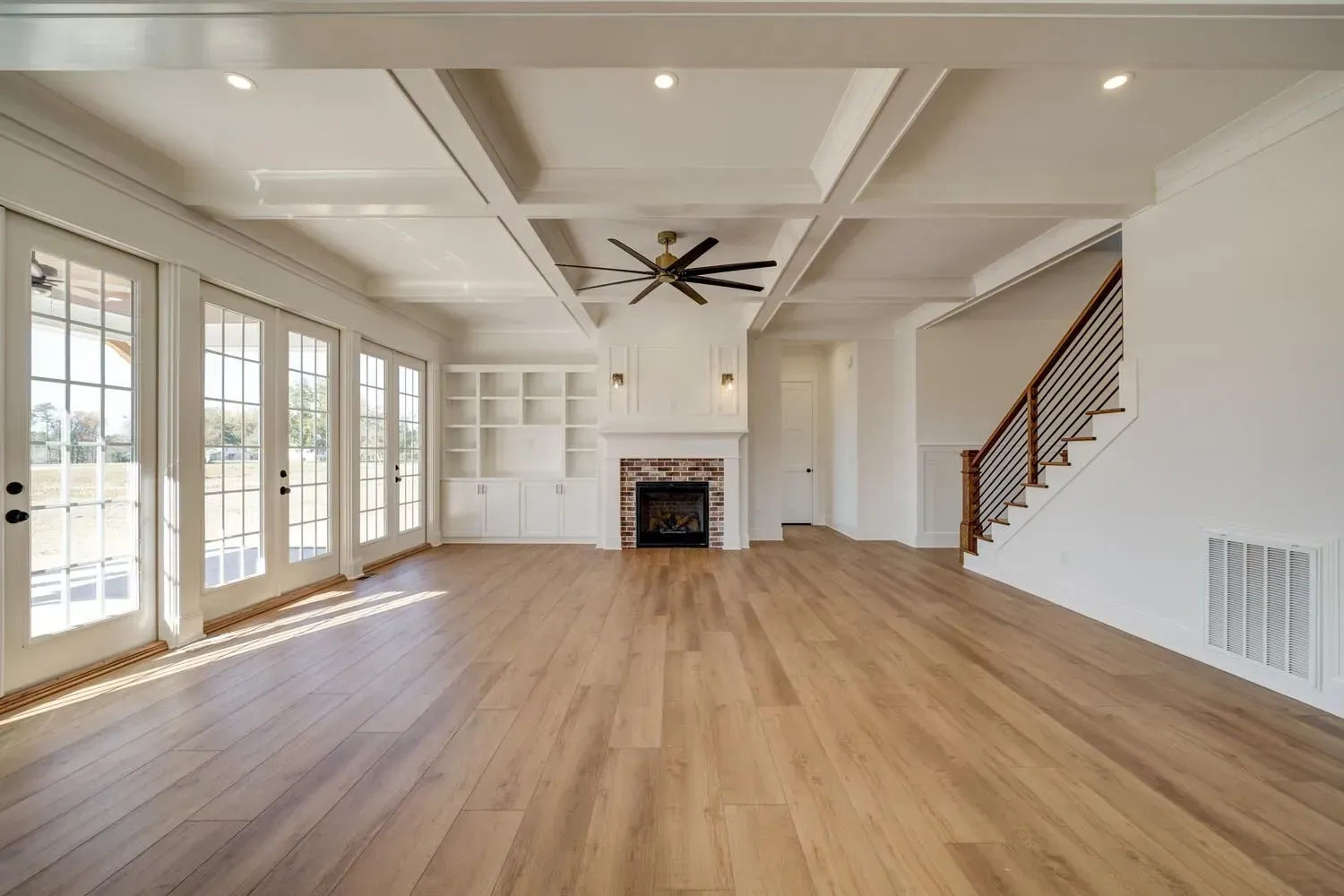 Bright, open-concept living room with light wood floors, a coffered ceiling, built-in shelving, a fireplace, and stairs.