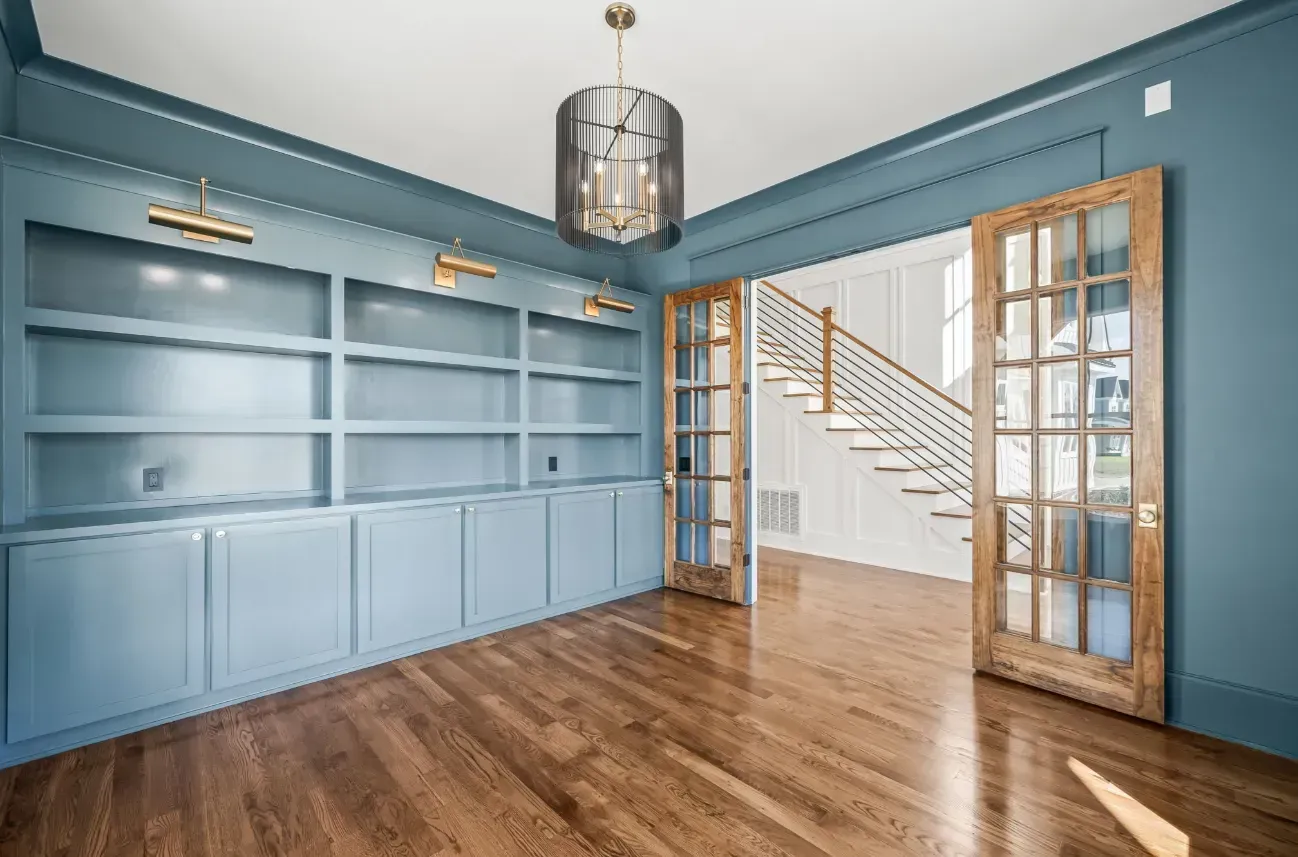 A home office with blue built-in shelving, hardwood floors, a pendant light, and glass-paneled wooden doors.