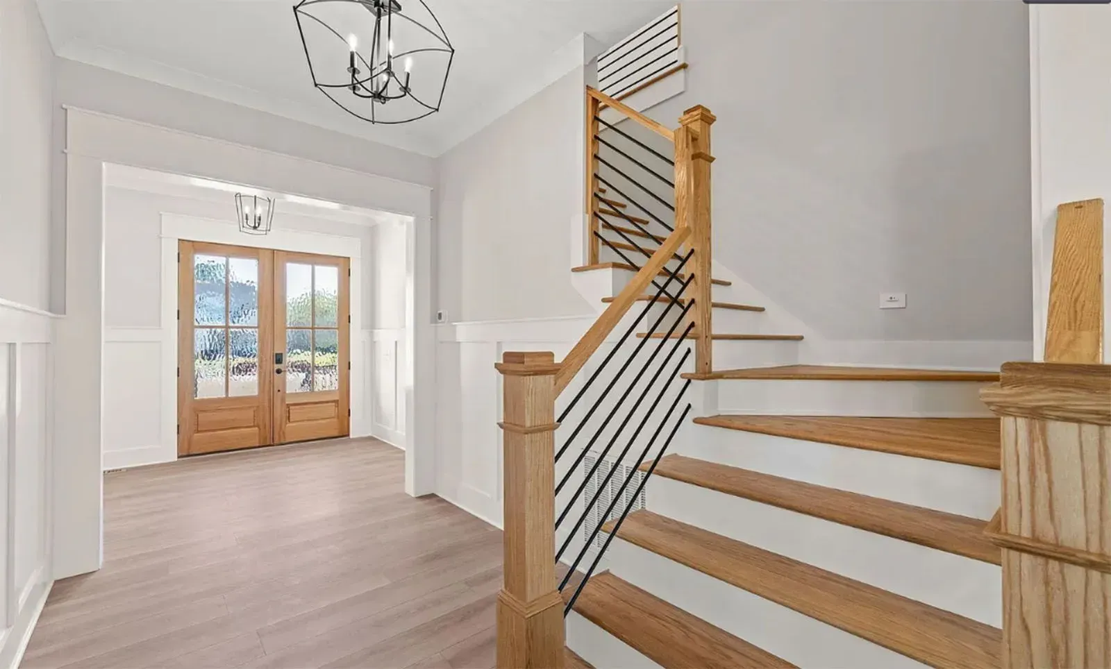 A bright home entryway with wood flooring, light walls, and a staircase featuring a wooden banister with iron spindles.