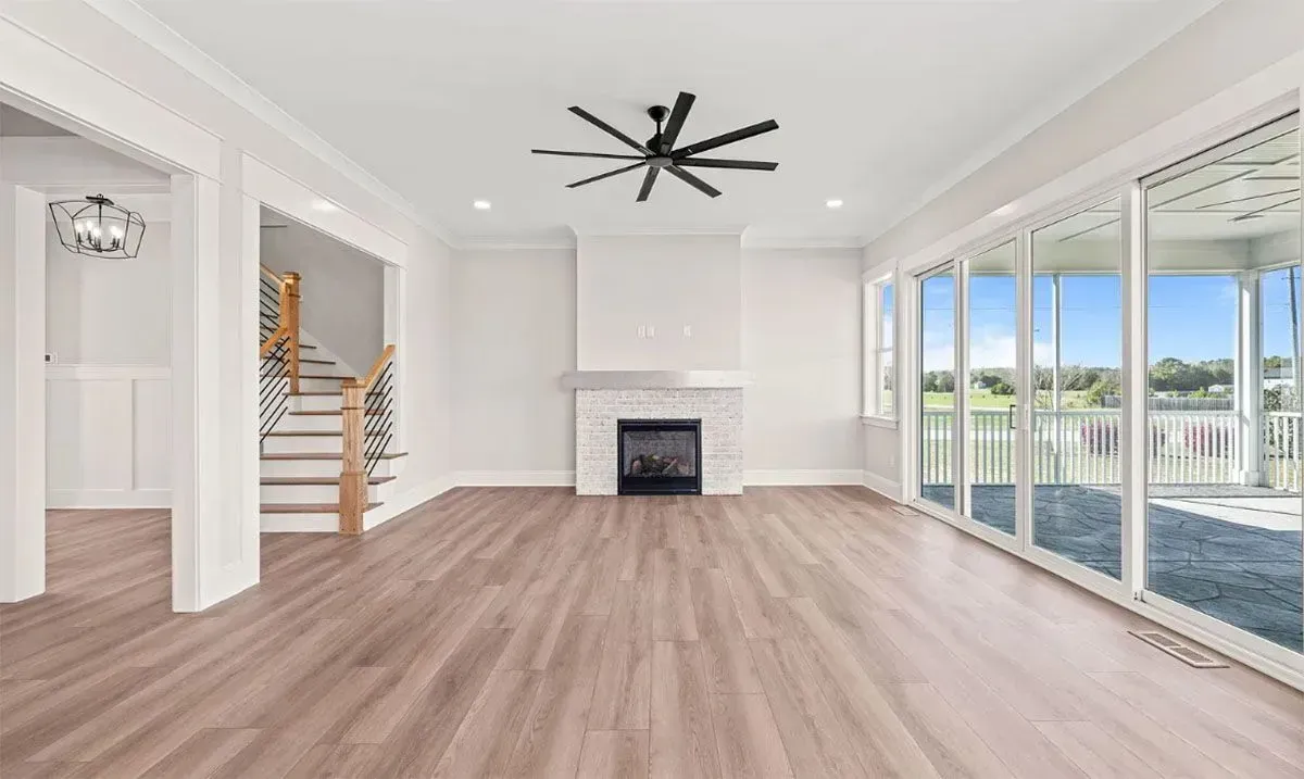 A bright, open-plan living room with light wood floors, a central fireplace, a ceiling fan, and large sliding glass doors.