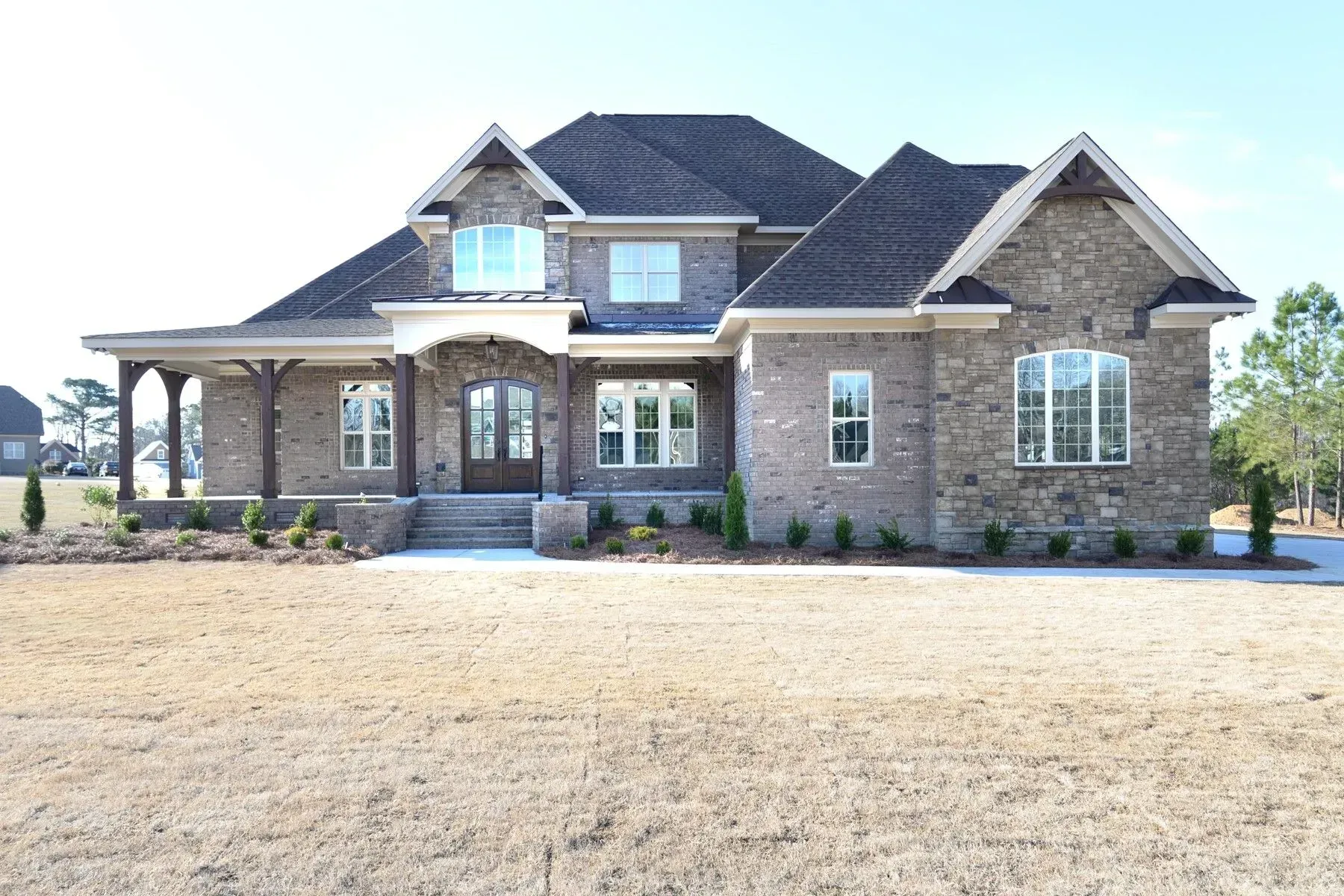 A two-story house with dark shingled roofs, stone and brick facade, a front porch, and a large, grassy yard.