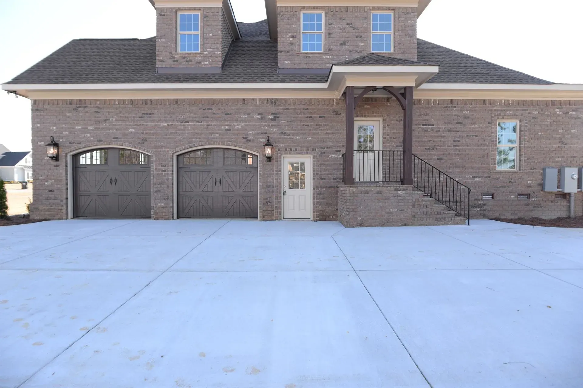A tan brick house with two garage doors, a small porch with stairs, and a wide concrete driveway.