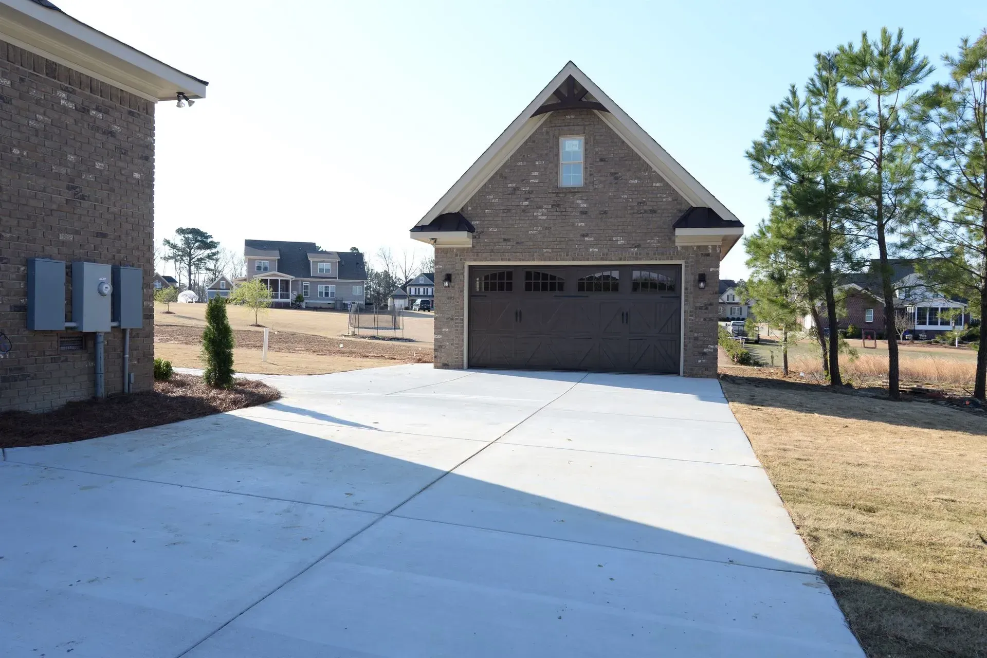 A concrete driveway leads to a brick detached garage on a sunny day in a residential neighborhood.