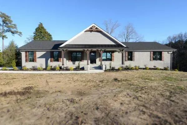 A tan, single-story house with a covered front porch, dark gray roof, and a large, grassy yard under a clear blue sky.