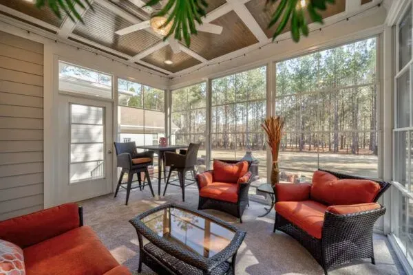 A screened-in porch with orange patio furniture, a glass coffee table, and a small dining set, overlooking a wooded yard.
