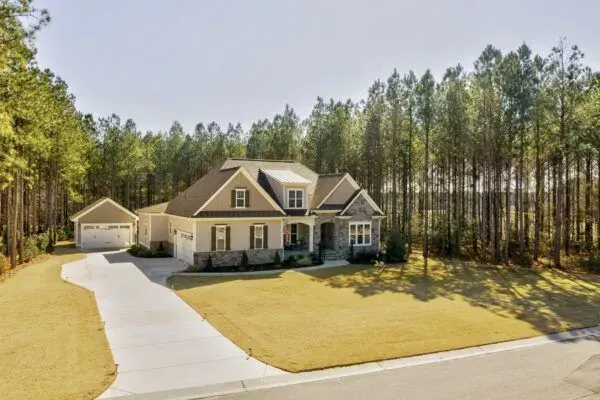 A two-story suburban house with a detached garage, tan siding, and stone accents, set against a backdrop of pine trees.