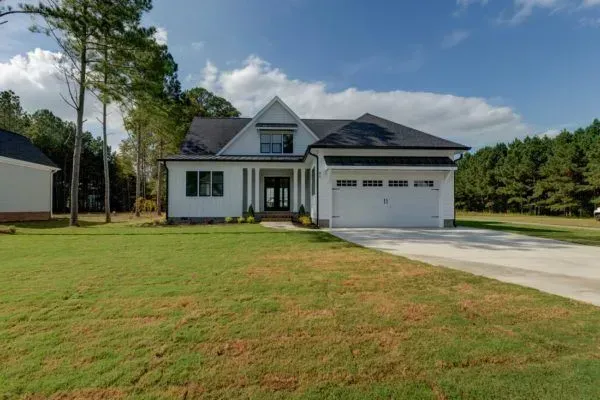 A white, two-story house with a black roof and attached garage, situated on a green lawn surrounded by pine trees.