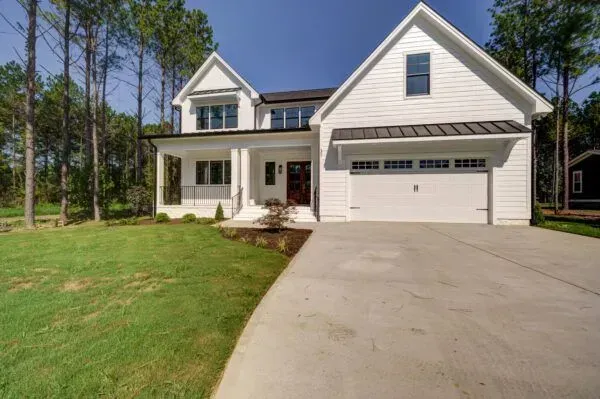 A two-story white house with a metal-roofed porch, garage, and a large concrete driveway in a wooded, sunny setting.