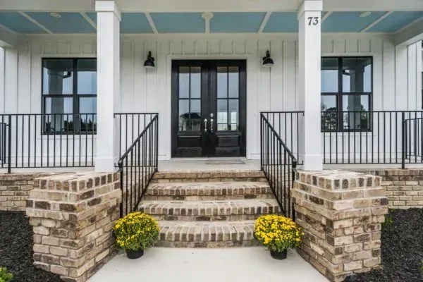 A front porch entrance with white vertical siding, black double doors, brick steps and pillars, and yellow potted mums.