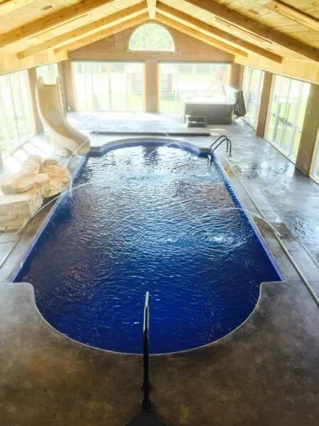 An indoor pool room featuring a dark blue pool, a curved slide, and a wooden vaulted ceiling with glass walls.