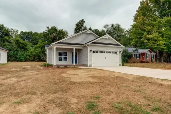 A light gray single-story suburban house with a white garage and covered front porch, set on a grassy lot with trees.