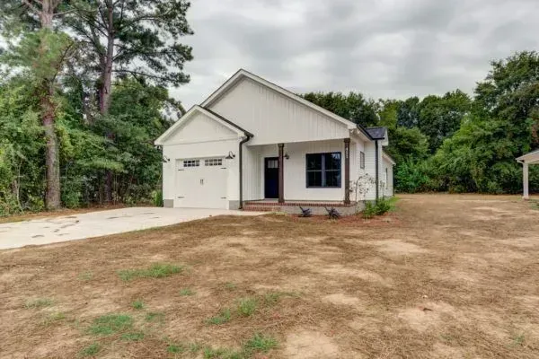 A white, one-story house with a single-car garage and a front porch, situated on a grassy lot surrounded by trees.