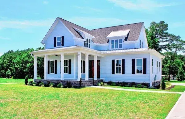 A two-story, light blue farmhouse with a front porch, dark shutters, and a dark roof, surrounded by a green lawn.