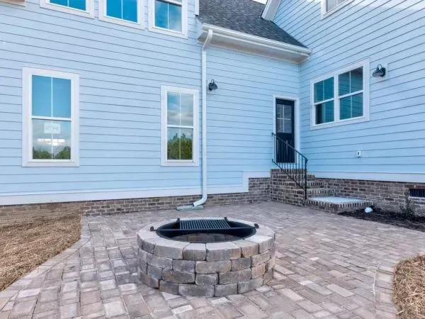 A stone fire pit sits on a paved patio outside a light blue house with a stone foundation and steps leading to a door.