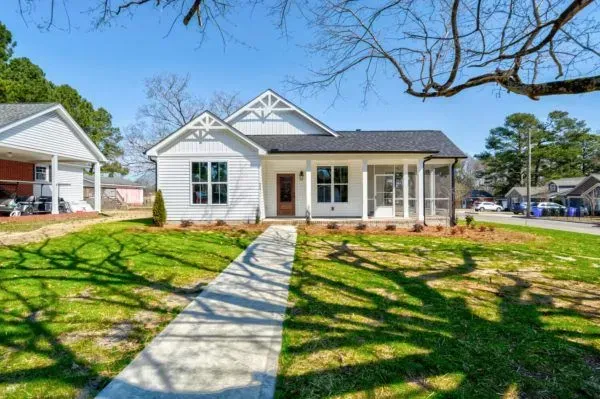 A white single-story cottage with a dark roof and front porch, viewed from a sidewalk across a sunny, tree-shaded lawn.
