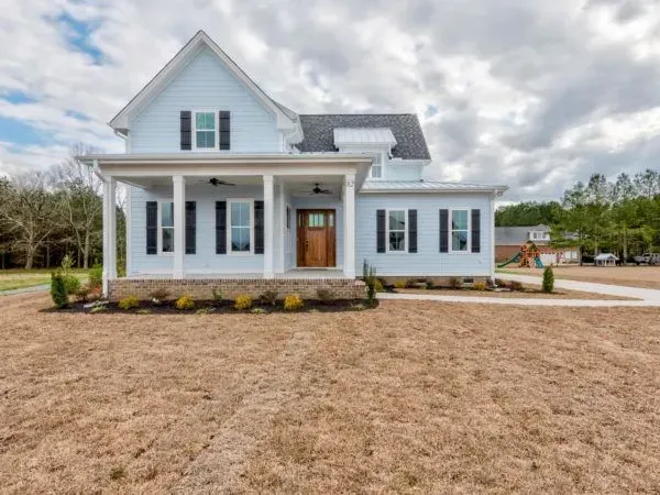 A light blue two-story house with a wooden front door, white porch columns, and black shutters, set on a grassy lawn.