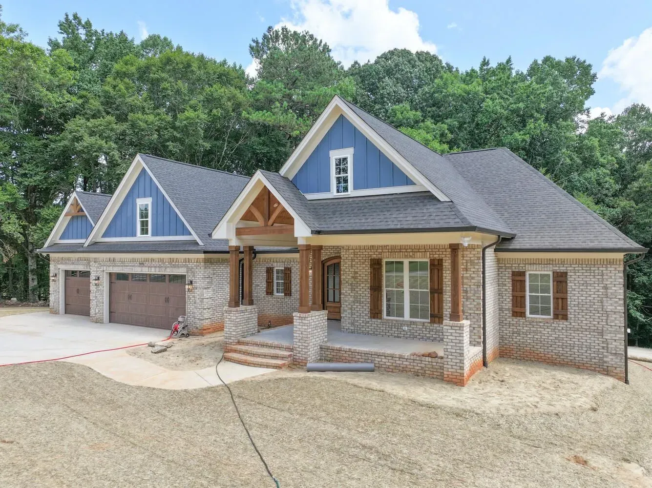 A single-story brick house with a blue-gabled exterior, covered porch, and three-car garage set against a wooded backdrop.