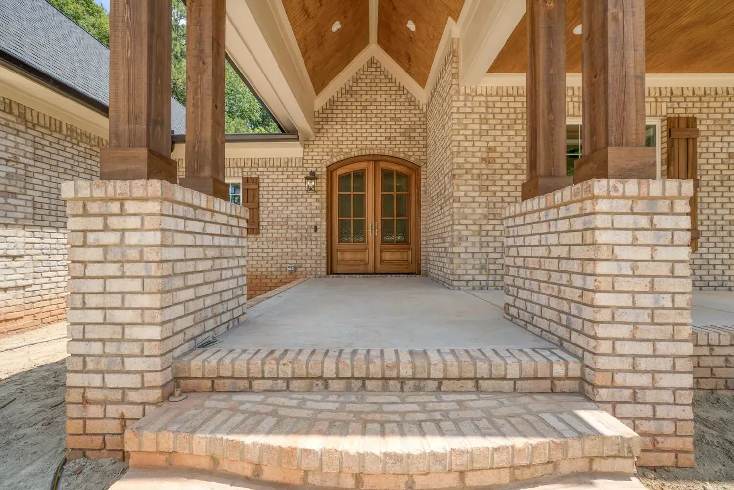 Entrance of a house with light brick siding, wooden columns on brick pedestals, and a double wooden arched door.