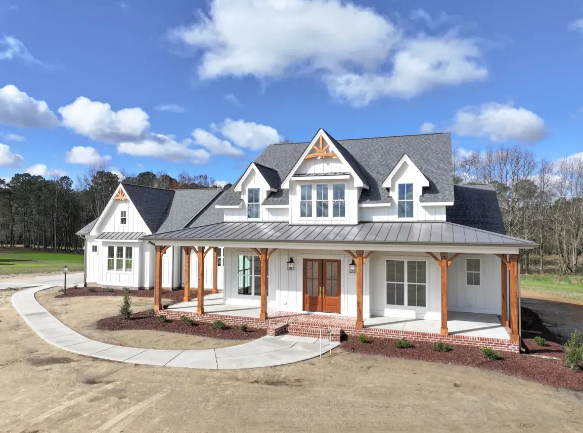 A white farmhouse-style house with a gray roof and covered wooden front porch, surrounded by a lawn and gravel path.