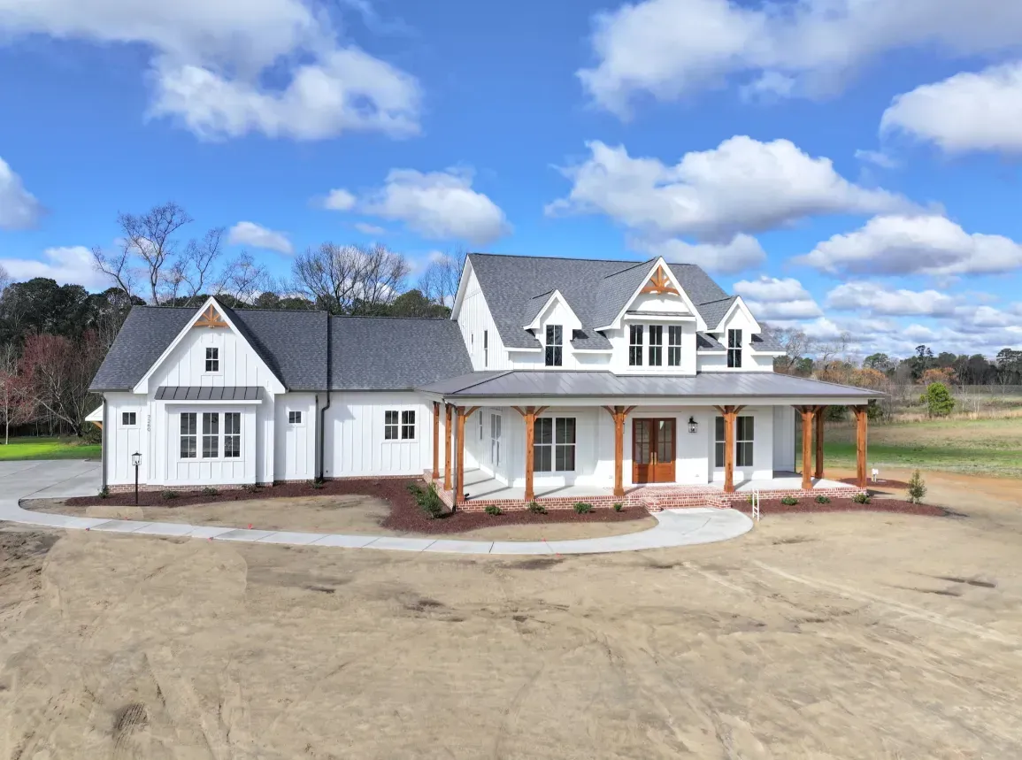 A white farmhouse with a grey roof, wood-post porch, and a wrap-around driveway against a blue, cloudy sky.