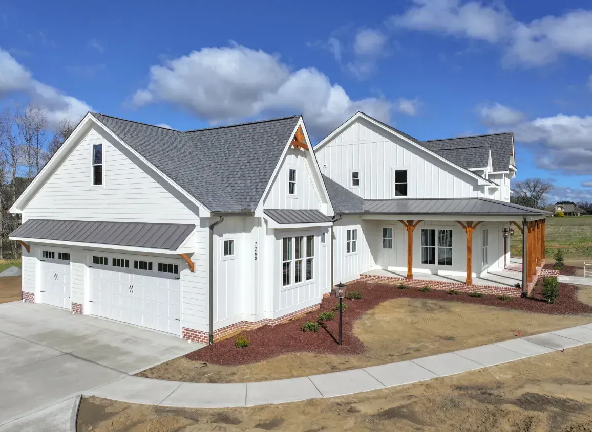 A modern white farmhouse with a metal roof, attached two-car garage, and a covered porch on a sunny, grassy lot.