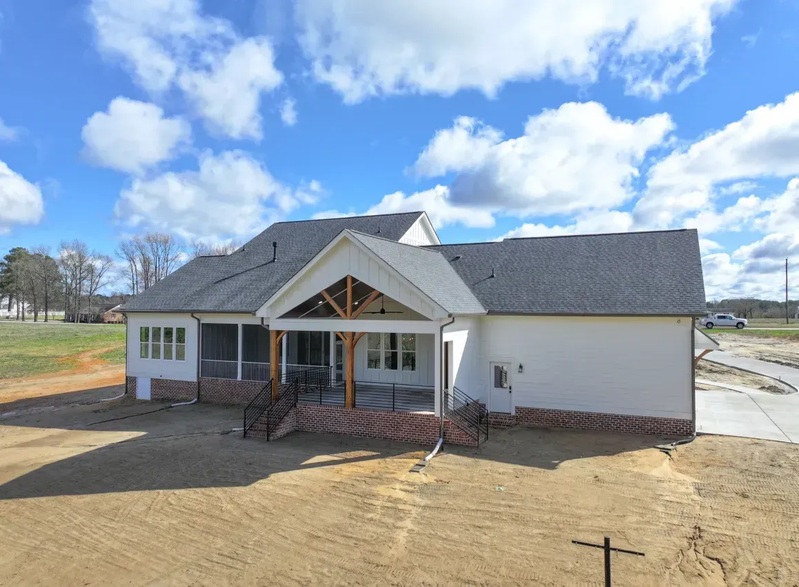 A white suburban house under construction with a wooden covered patio and dark gray roof against a blue, cloudy sky.