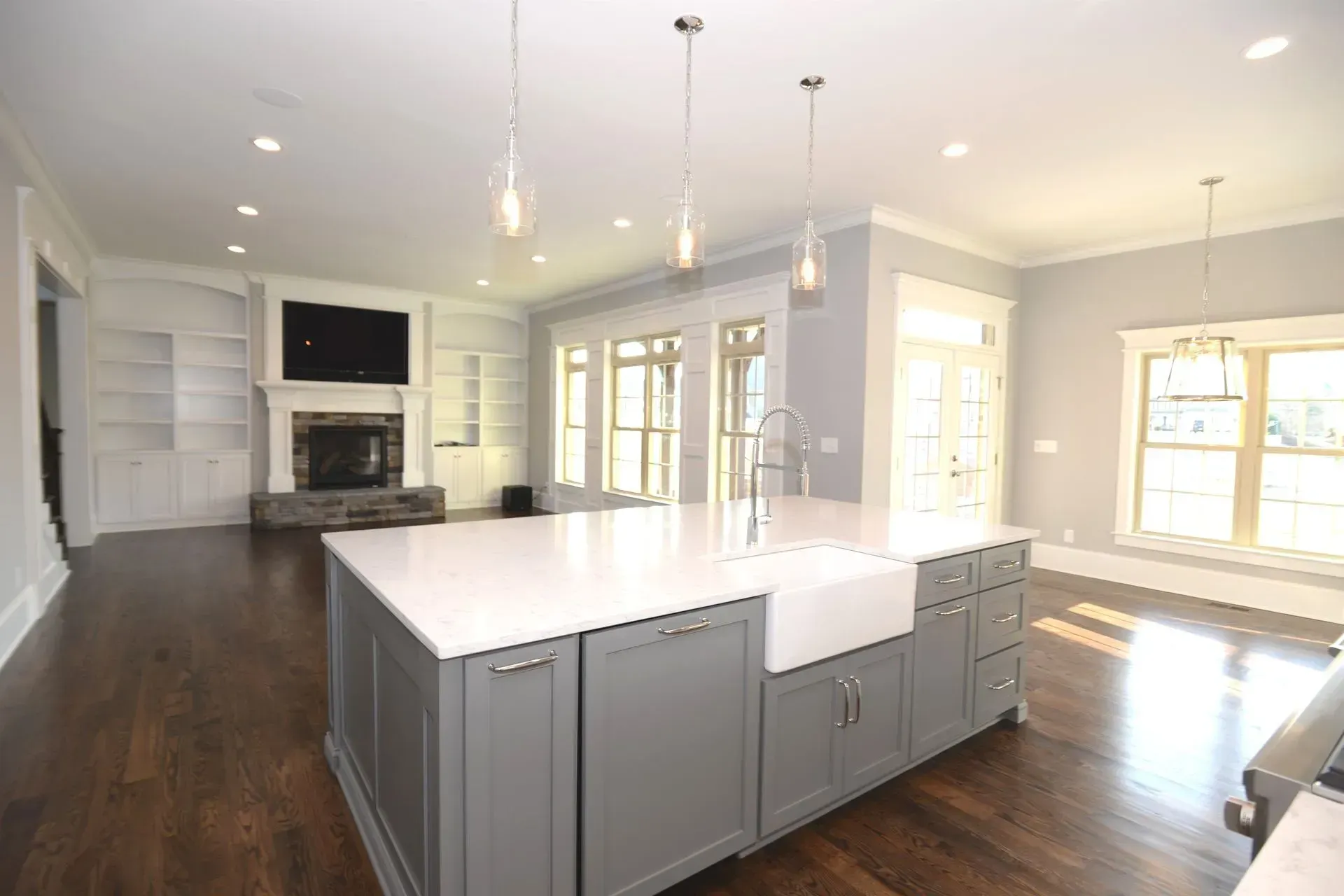 A kitchen featuring a large gray island with a white farmhouse sink, hardwood floors, and a living area with a fireplace.