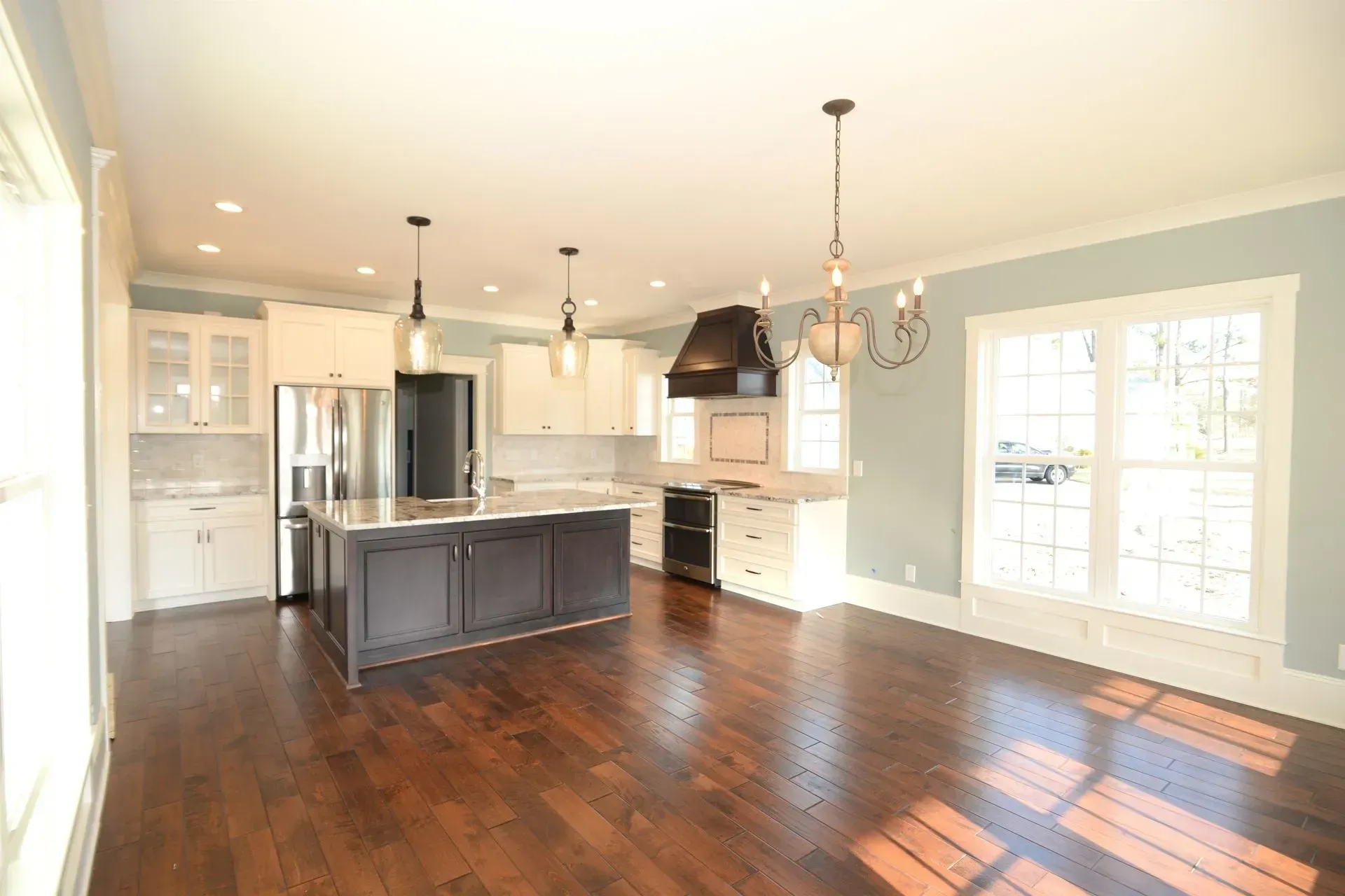 A bright kitchen with white cabinets, a dark island, stainless appliances, a black range hood, and dark hardwood floors.