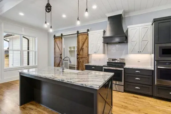 A modern farmhouse-style kitchen with a central island, dark cabinetry, white upper cabinets, and wood barn doors.