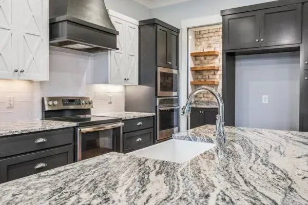 A modern kitchen featuring white and charcoal cabinets, granite countertops, a farmhouse sink, and a brick-walled pantry.