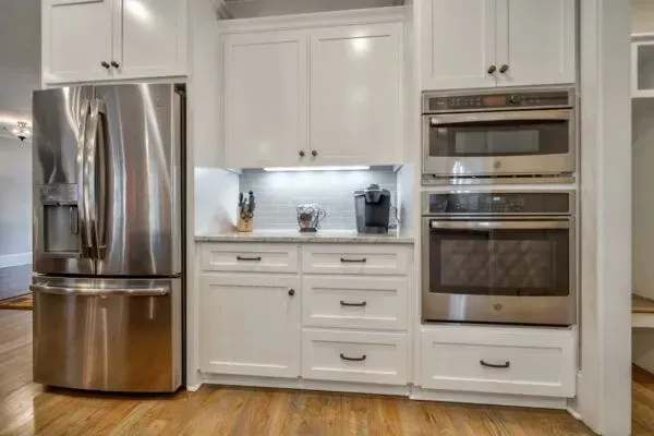 Stainless steel refrigerator and double wall oven flanking a white kitchen cabinet unit with drawers and appliances.