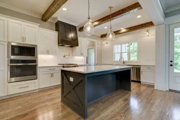 Modern white kitchen with a dark central island, wood ceiling beams, stainless steel appliances, and hardwood floors.