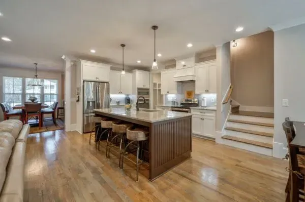 A modern kitchen featuring a dark wooden island with bar stools, white cabinetry, hardwood floors, and a nearby staircase.