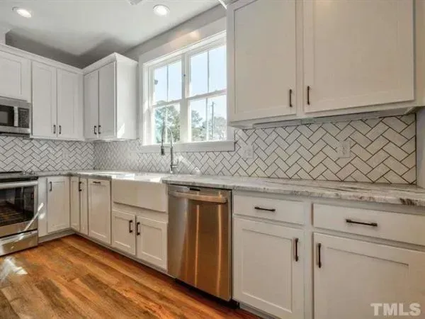 A bright kitchen with white shaker cabinets, stainless steel appliances, marble countertops, and herringbone tile backsplash.