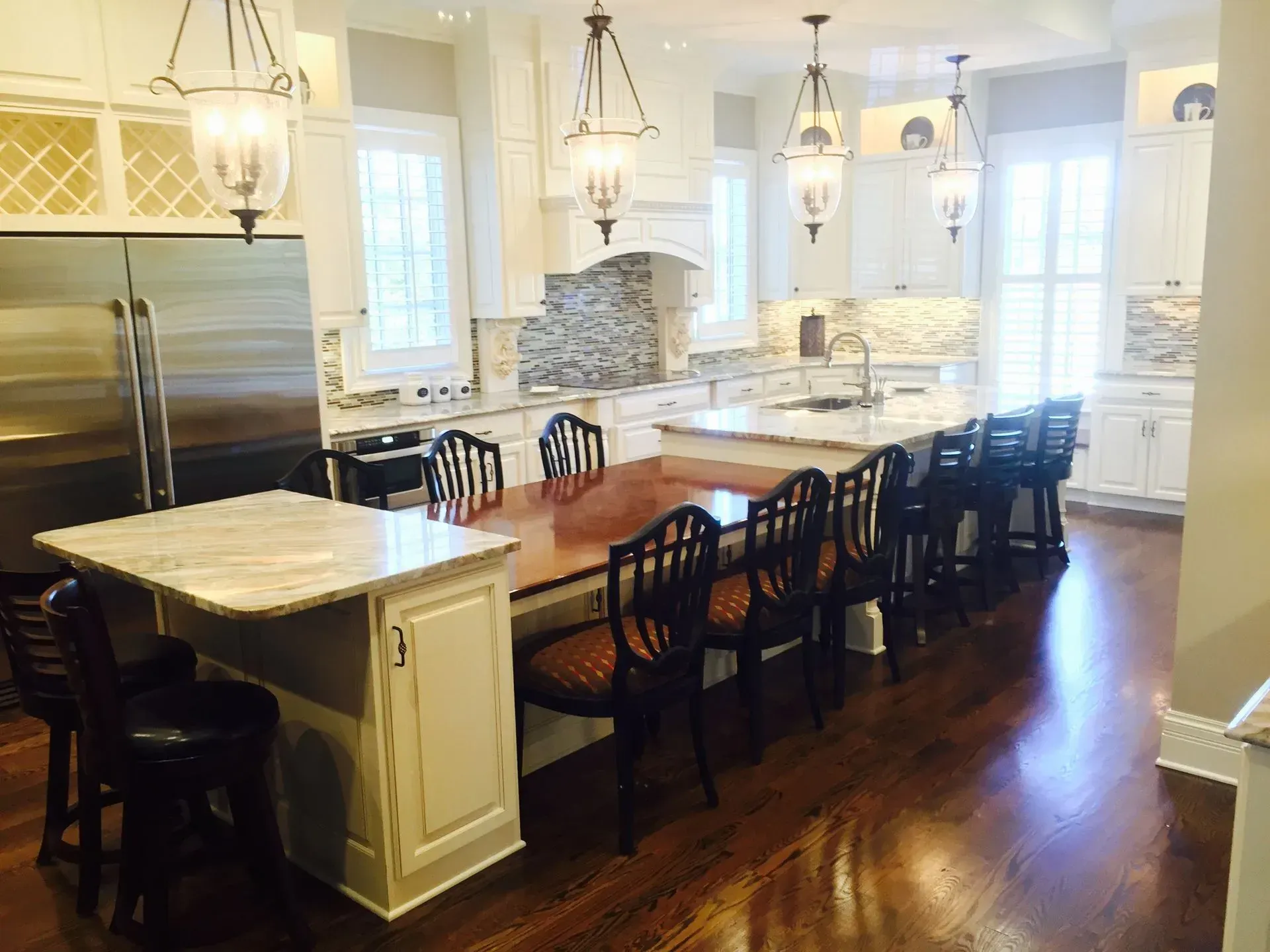A bright, white kitchen with a large island, wooden dining table, black chairs, glass pendant lights, and hardwood floors.