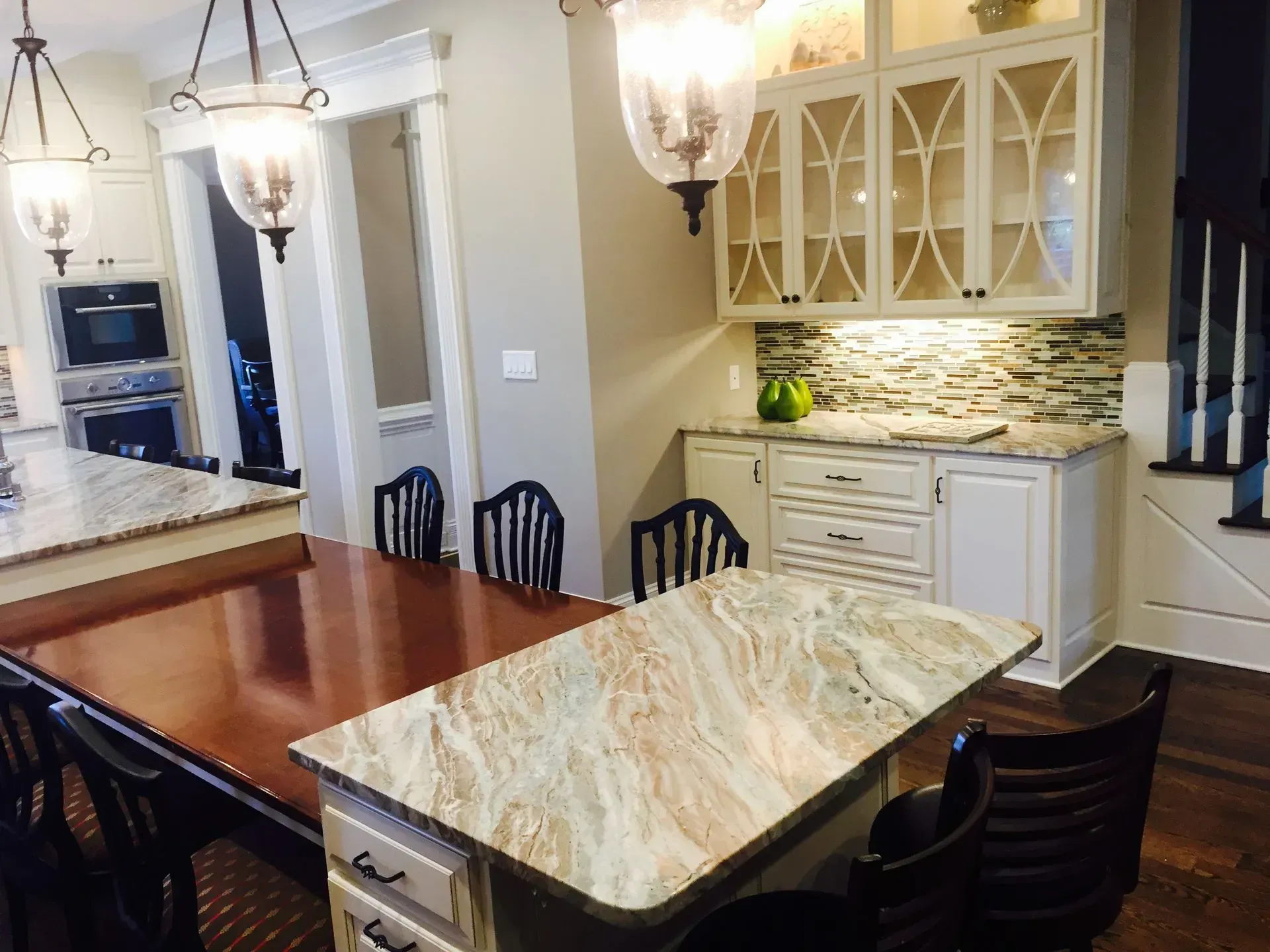 A kitchen interior with a marble-topped island, dark wooden stools, white cabinets, glass display doors, and warm lighting.